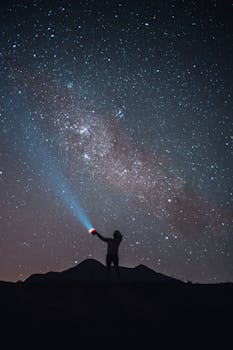 在无云的星空下，一名匿名游客的剪影站在悬崖上，用手电筒发出信号