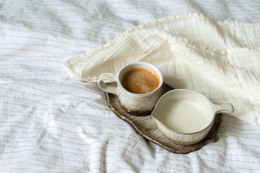 A cozy scene with a ceramic cup of coffee and milk on a textured plate on bed linen.
