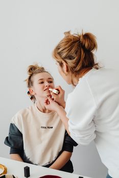 A mother applies lipstick to her daughter, showcasing a bonding moment in a well-lit room.