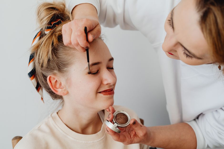 Teenager smiling while receiving makeup application from a woman indoors.