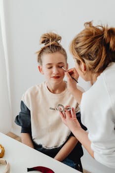 A mother applies eyeshadow to her daughter's closed eyes in a casual home setting.