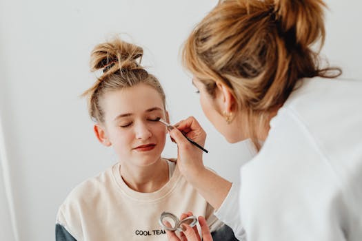 A makeup artist skillfully applies eyeshadow on a teenager with a relaxed expression.