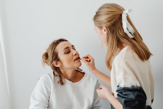 A young woman applies lipstick to another in a well-lit indoor setting.