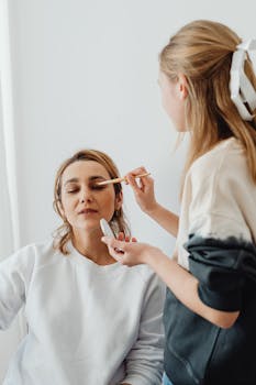 A woman getting makeup applied by a friend indoors. Bright, casual setting.