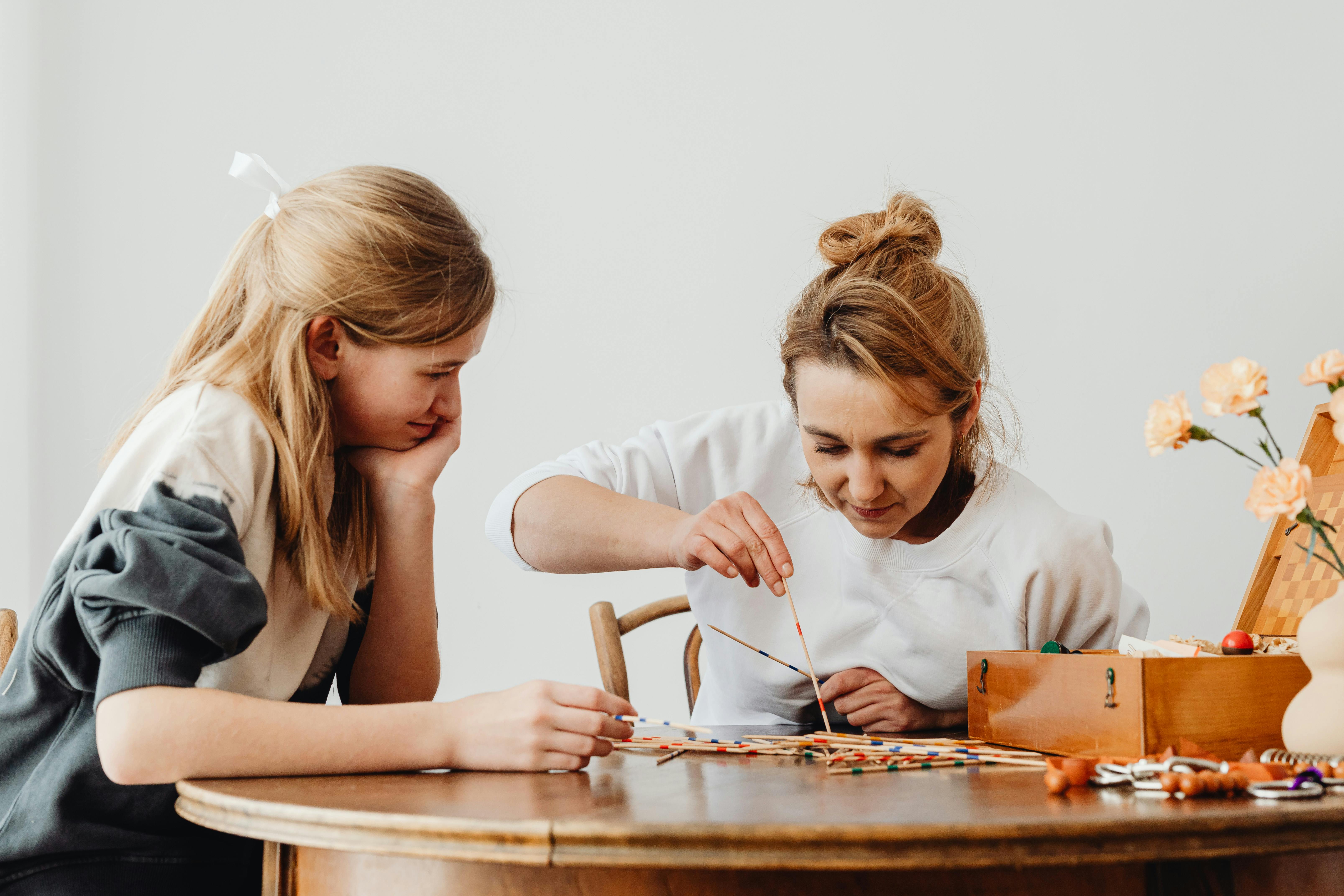 A mother and daughter sitting indoors engage in a creative painting activity at a wooden table.