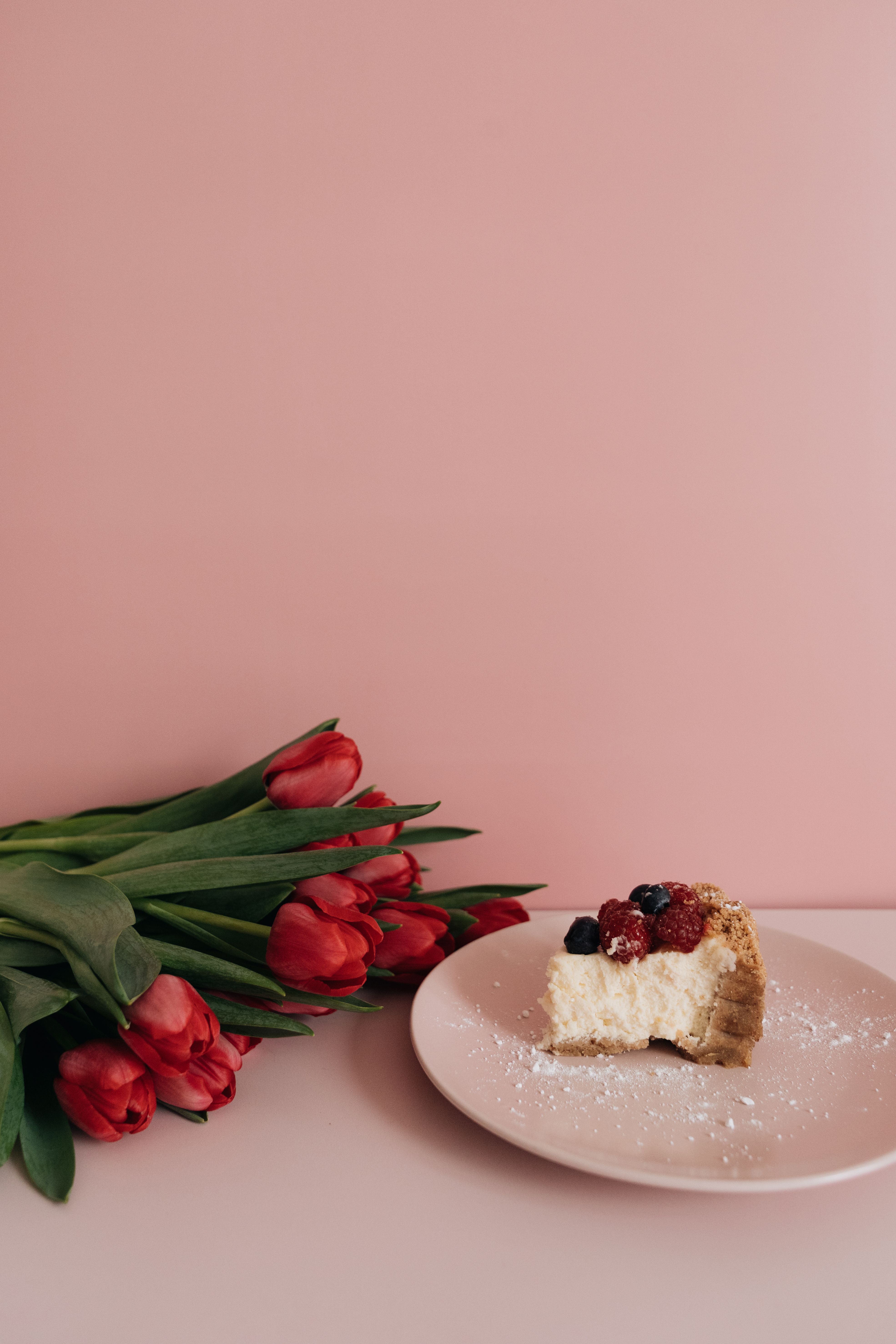 A delicious slice of cheesecake with berries and a bouquet of red tulips on a pastel pink background.