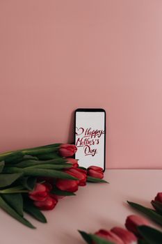 Red tulips and smartphone with Mother's Day greeting on pink background.