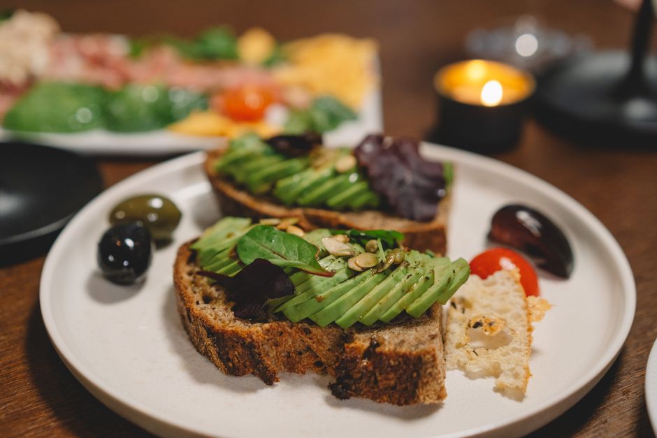 A toasted slice of seeded whole grain bread topped with mashed avocado, flaky salt, and red pepper flakes, with the sliced loaf and a few extra sunflower seeds scattered on a wood board behind it.