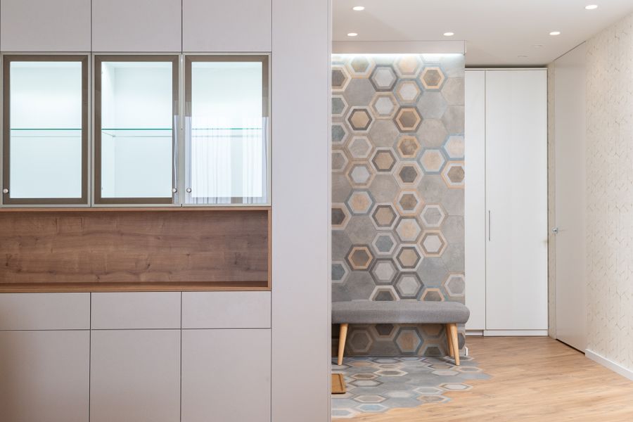 Glass cabinets in white kitchen against modern hallway with fabric bench and wall with hexagonal ornament