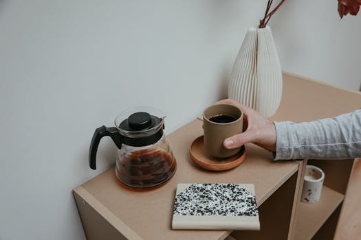 Aesthetic coffee scene with ceramic cup, teapot, notebook, and vase on a tabletop.