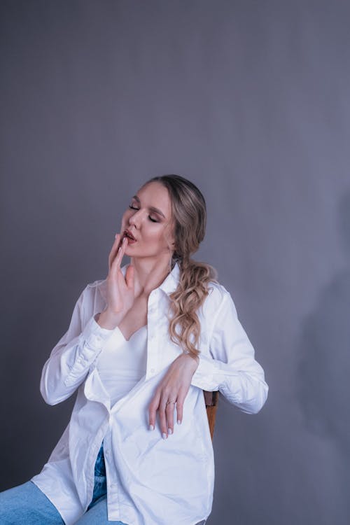 Thoughtful young woman in shirt sitting on chair with closed eyes and hand on mouth on gray background in bright studio