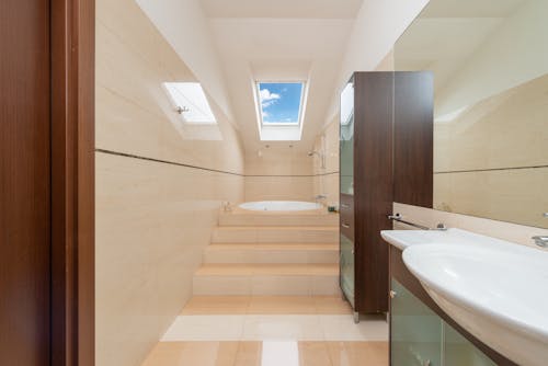 Modern bathroom interior with washbasin above cabinet on tiled floor with staircase and bath under window at home