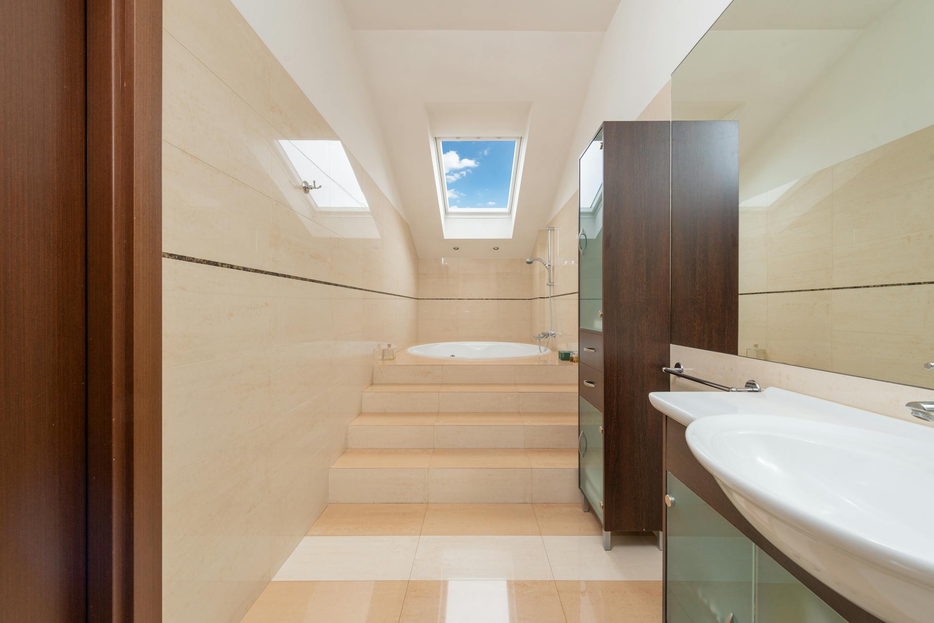 Modern bathroom interior with washbasin above cabinet on tiled floor with staircase and bath under window at home