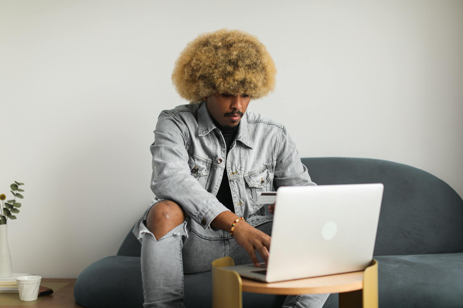 Young man with afro hairstyle shopping online using laptop and debit card in a modern, relaxed home setting.