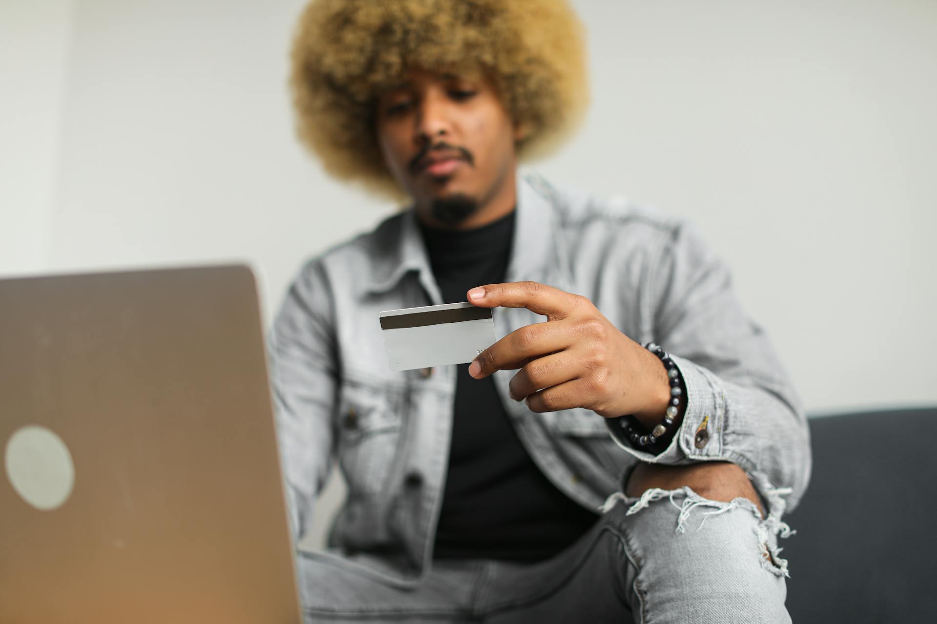 A man with afro hair using a credit card for online shopping while sitting indoors with a laptop.