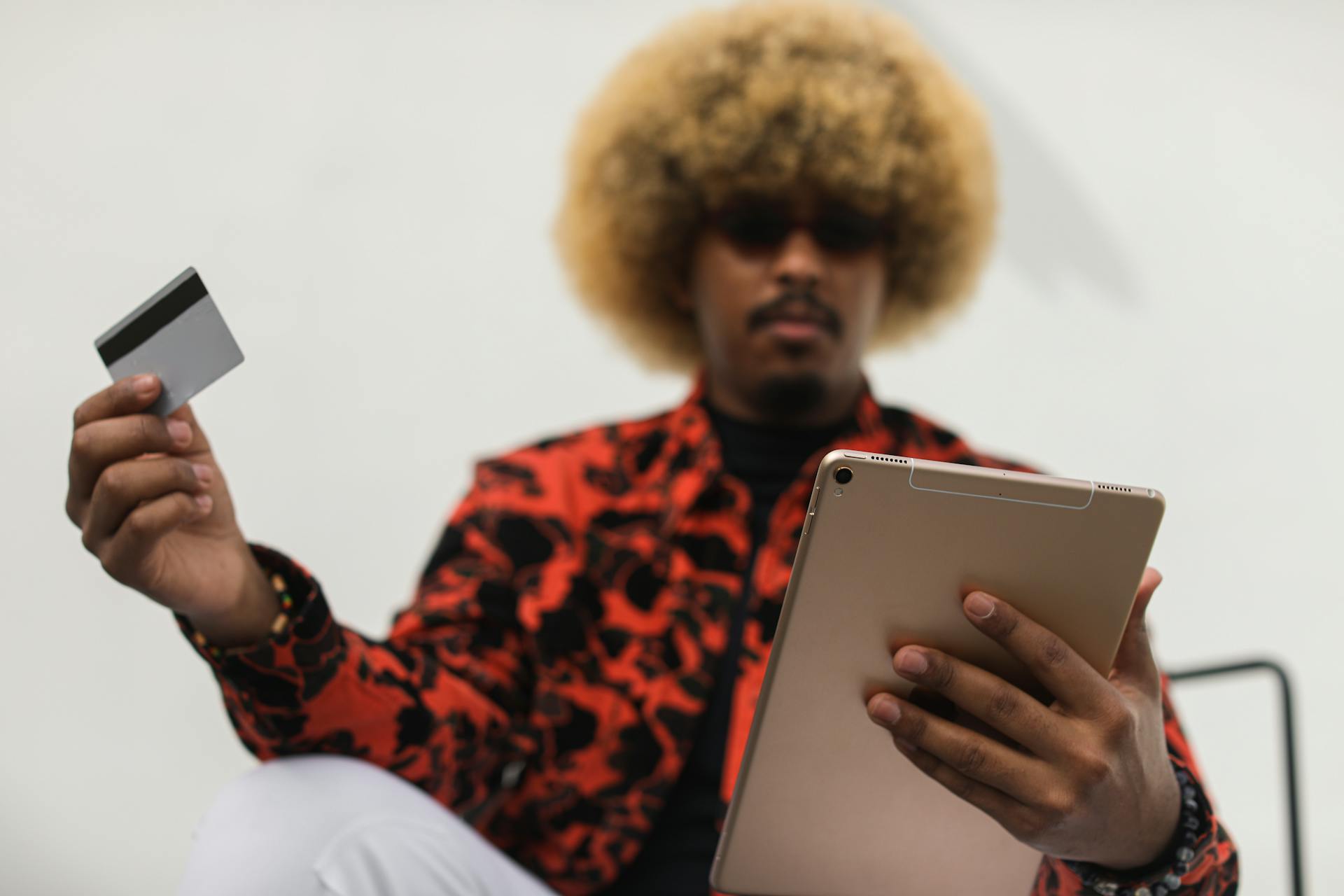 Fashionable individual with afro hair using a tablet and credit card for online shopping indoors.