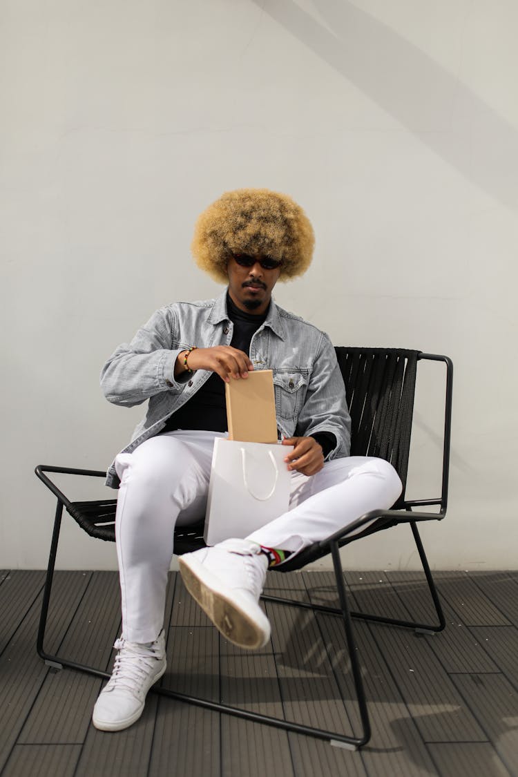 Man With Blonde Afro Hair Sitting On A Chair Holding A Box From A Paper Bag