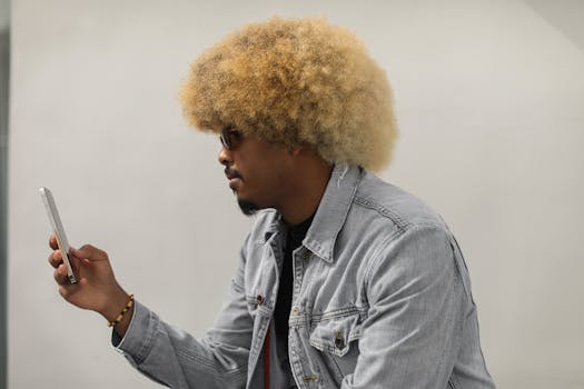 Side-profile of a man with afro hair wearing sunglasses and denim jacket, holding a smartphone.