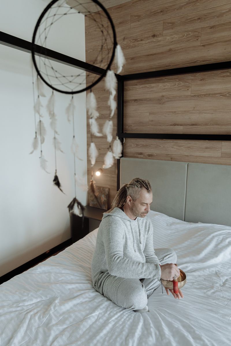 Meditative scene with a man seated on a bed, holding a meditation bowl, under a dreamcatcher.