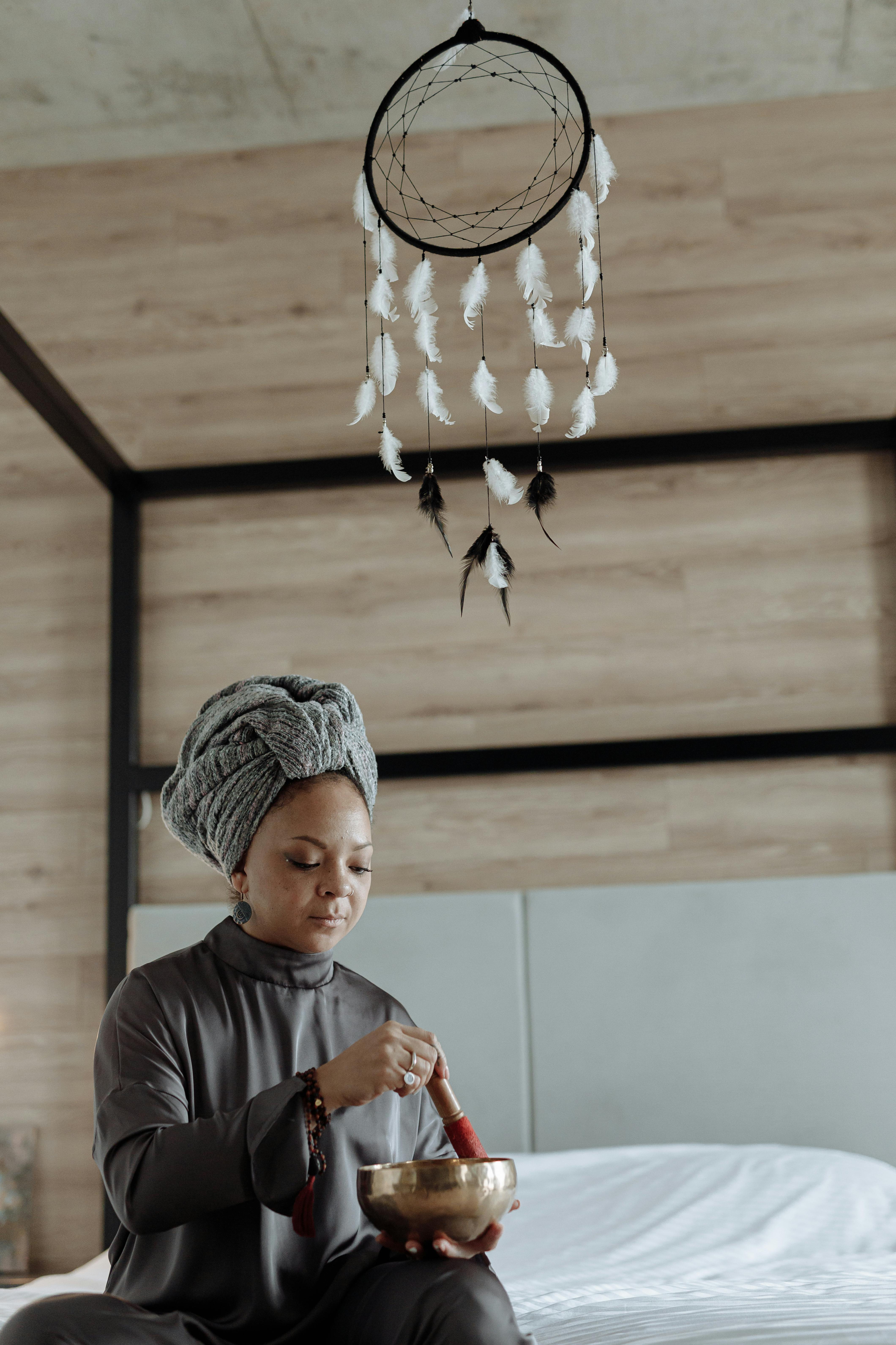 Woman meditating indoors with a singing bowl beneath a dreamcatcher, promoting tranquility and mindfulness.