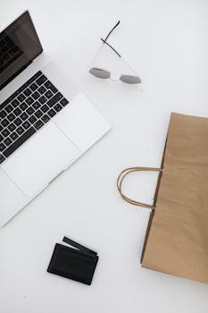 Top-down view of a minimalist desk setup featuring a laptop, sunglasses, wallet, and paper bag.