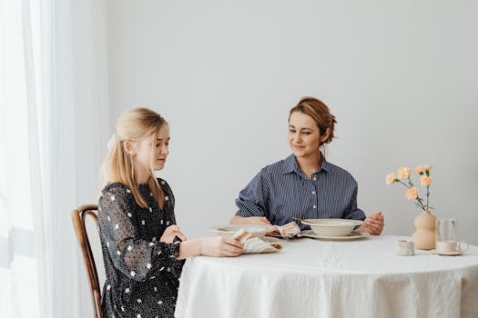 A mother and daughter sit at a table with flowers, enjoying dinner in a cozy indoor setting.