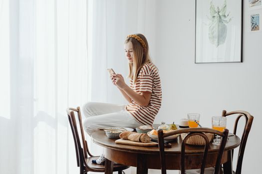 Woman sitting on a table using her phone, surrounded by a stylish breakfast setting in a bright dining room.