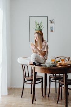 A young woman sitting on a dining table, using a smartphone in a bright room.