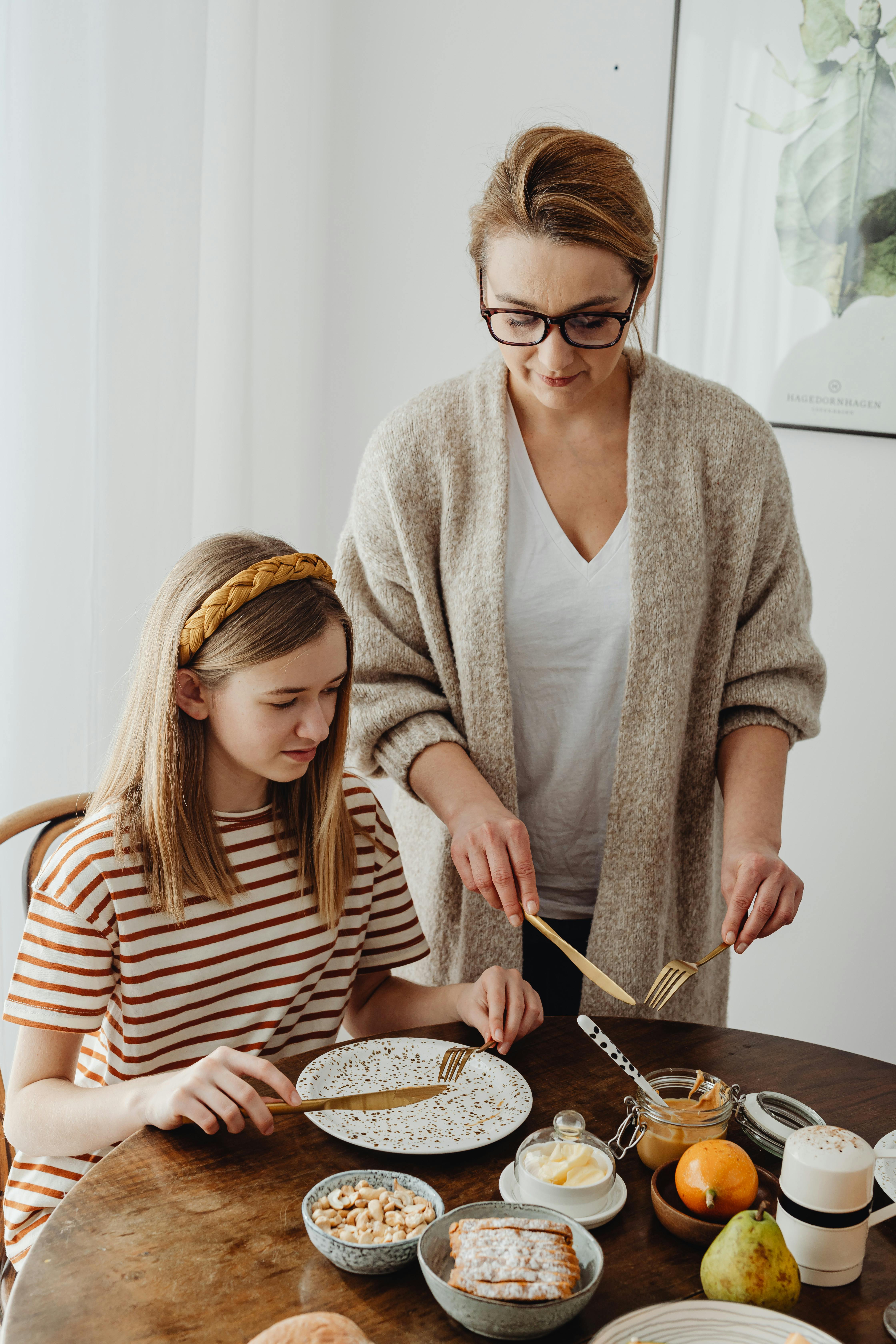 A woman guides a teenage girl on dining etiquette at a wooden table set with breakfast food.