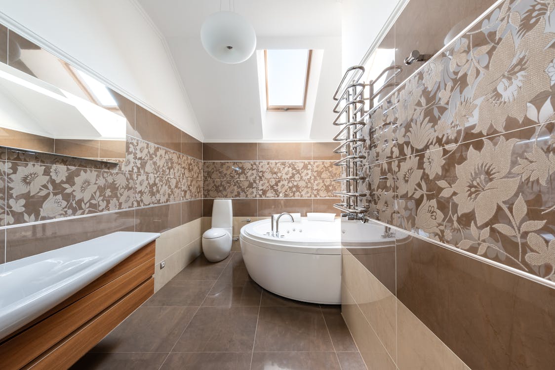 Round ceramic bathtub and toilet bowl in modern bathroom with brown tiled walls in house in sunlight