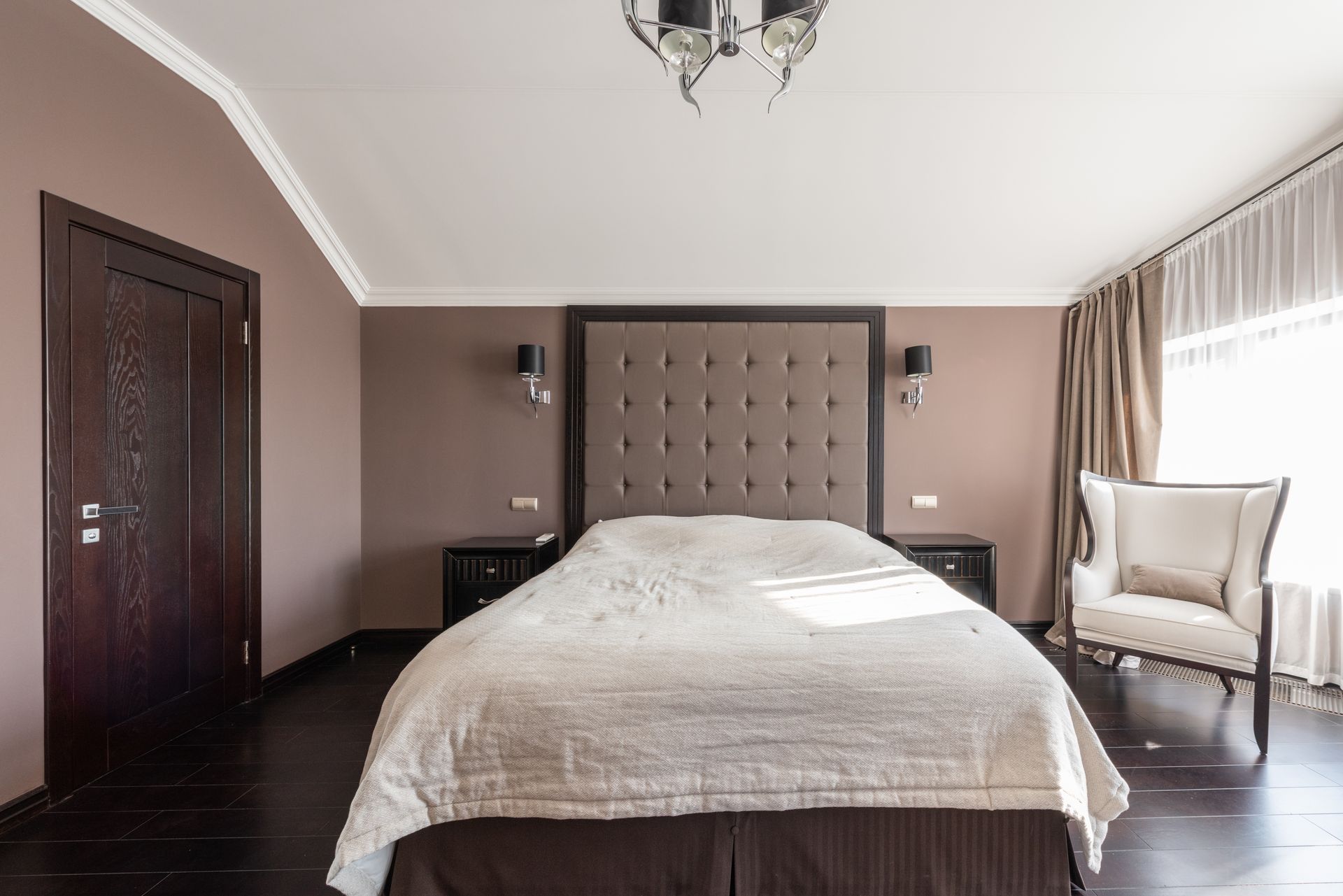 Comfortable bed and armchair in classic styled bedroom with wooden floor and door on sunny day