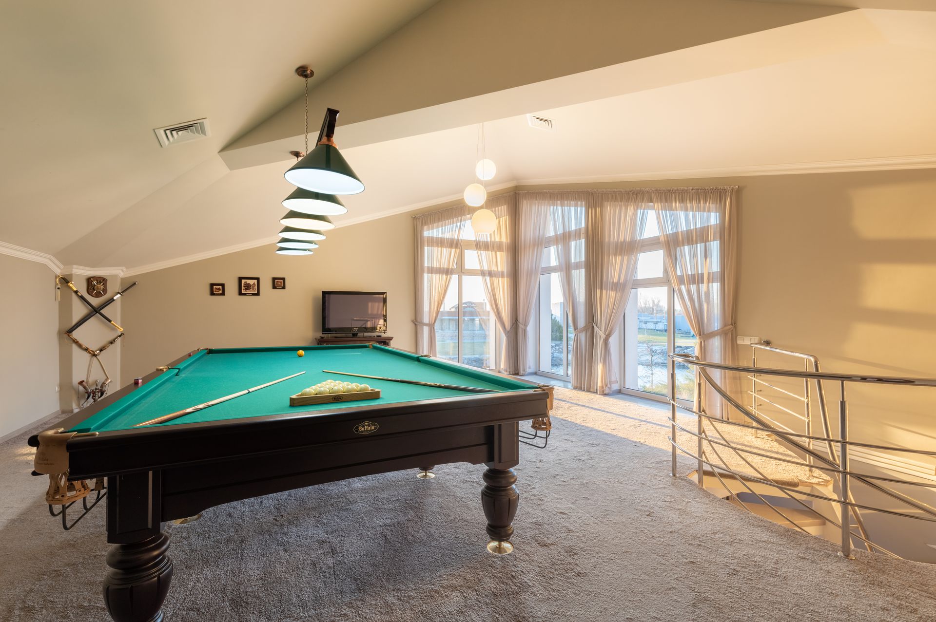Wooden pool table with cues placed on carpet in spacious room with windows and staircase with metal railing in contemporary building