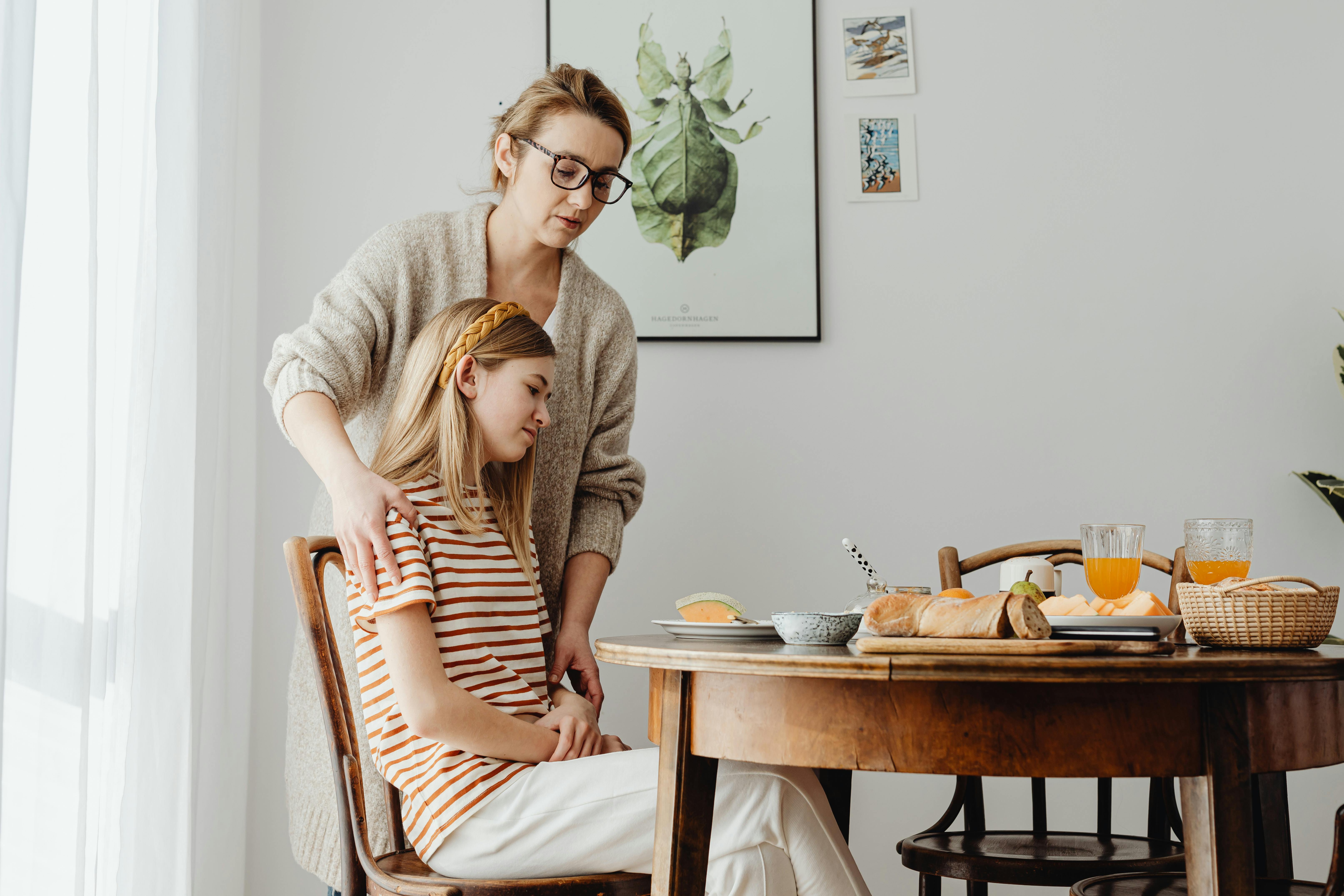 Free A mother comforts her teenage daughter during breakfast. A warm family moment at the dining table. Stock Photo