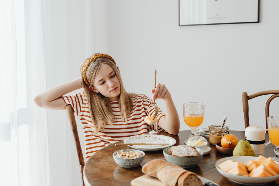 Teen girl in striped shirt at breakfast table with fruits, bread, and juice. Casual morning setting.