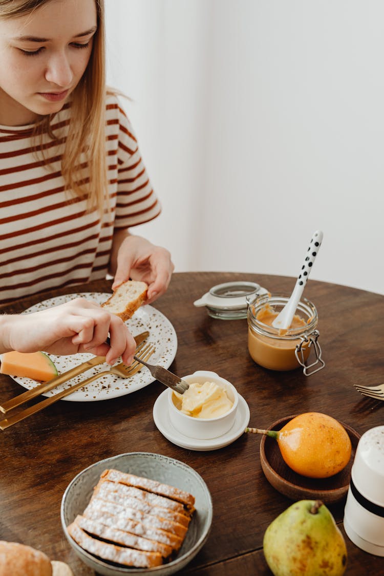 Young Woman Sitting At The Table And Spreading Butter On Bread 