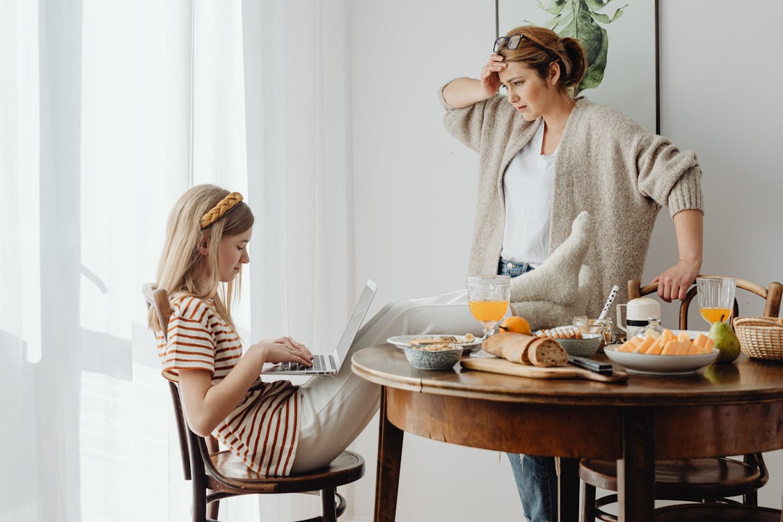 Free Mother Feeling Annoyed With Her Daughter With Feet On Table While Using Laptop Stock Photo
