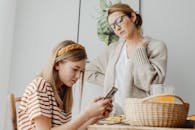 A mother discusses with her daughter engrossed in her phone during breakfast indoors.