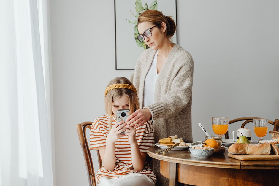 Mother assisting daughter with phone during breakfast. Cozy family morning at home.
