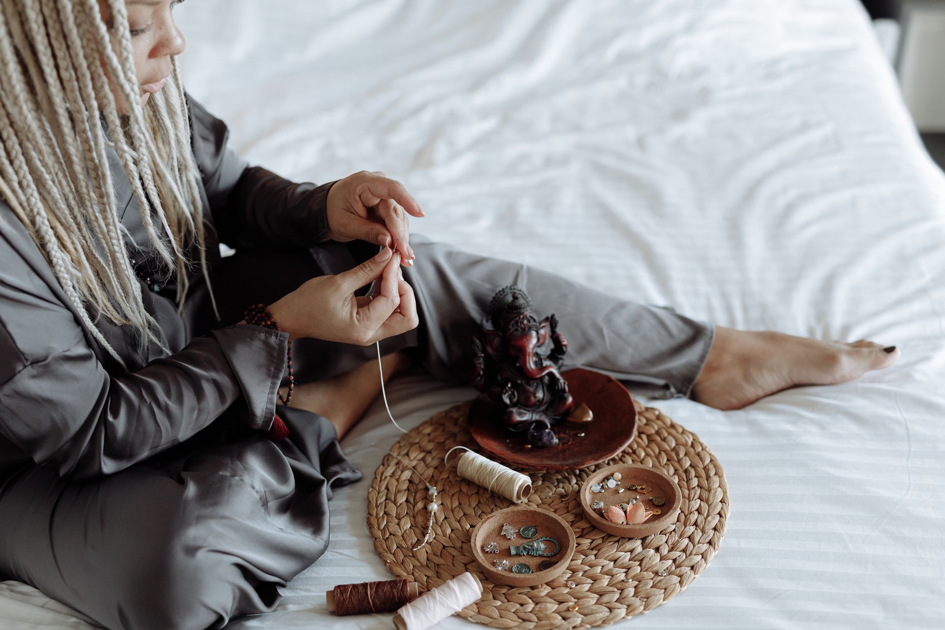 Woman with dreadlocks engaged in jewelry crafting using sewing threads and spools indoors.