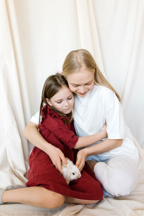 Free A mother and daughter lovingly embrace while holding their pet rabbit indoors. Stock Photo