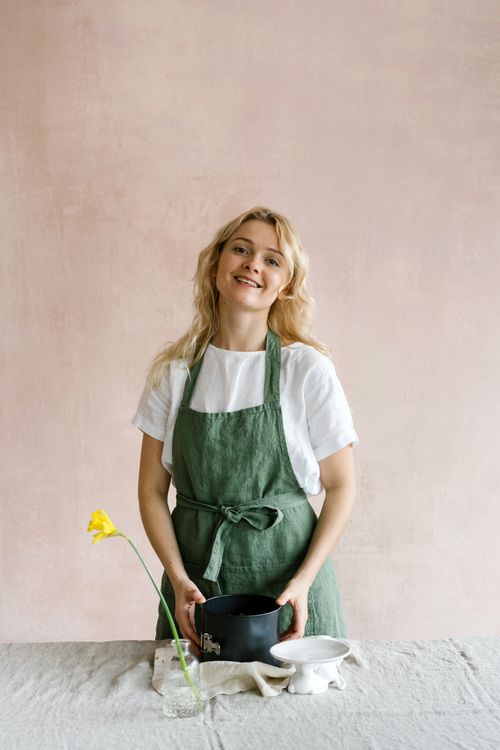 Free Charming baker smiling in a green apron with kitchen tools on a table indoors. Stock Photo