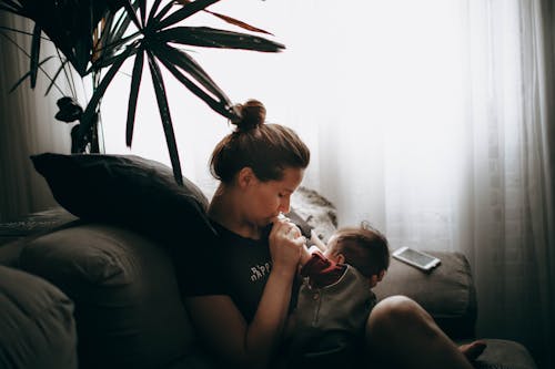 Side view of caring mother sitting on comfortable sofa with little son while cuddling in light living room at home