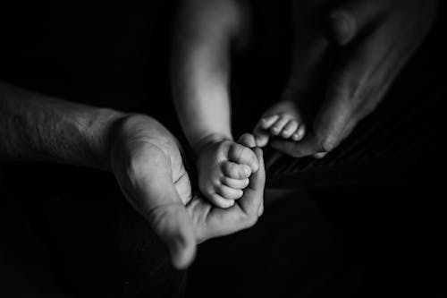 Black and white of caring unrecognizable father touching feet of anonymous little baby in arms while spending time at home