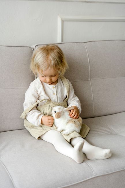 A blonde toddler in a dress sits on a sofa holding a plush bunny, exuding a cozy and heartwarming vibe.