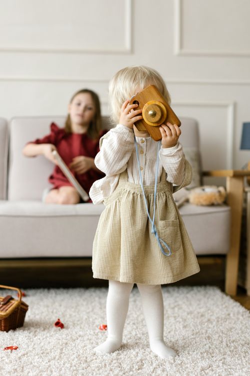 Free A young child plays with a wooden toy camera indoors while an older sibling reads on the couch. Stock Photo