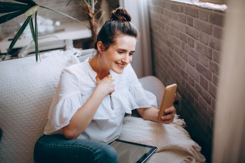 Free From above of young smiling female in casual clothes sitting on comfortable sofa with netbook and browsing smartphone while spending time at home Stock Photo