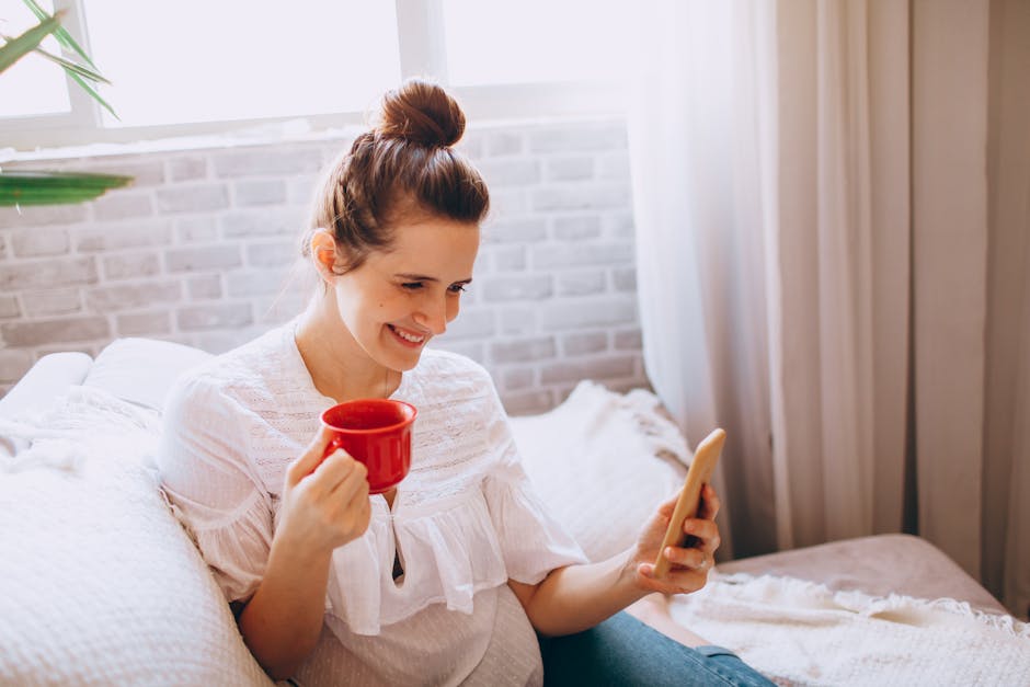 Young cheerful female in casual clothes sitting on sofa and drinking coffee while using smartphone