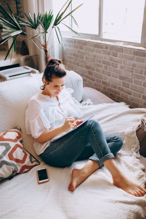 Free Young woman writing in notebook while sitting on sofa Stock Photo