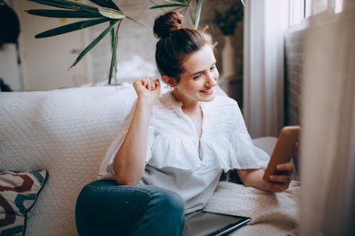 Free Smiling female in casual clothes sitting on comfortable sofa in light room and browsing smartphone Stock Photo