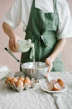 A person in a green apron pours milk into a bowl while preparing food with eggs on a table.
